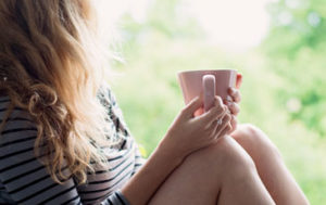 Kvinna som chillar, tittar ut genom fönstret och håller en rosa kaffekopp i handen. Vad är Mejvård för dej?